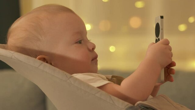 Funny kid watching TV show series cartoons holding remote control for TV digital media player, 9-month-old baby boy face closeup. High quality 4k footage