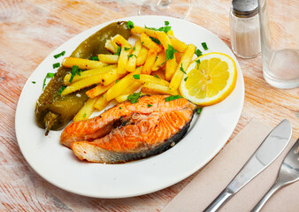 Salmon steak with fried potato, green pepper and slice of lemon served on white plate with serving pieces.