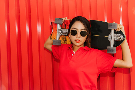 Portrait beautiful Asian hipster girl in red shirt with stylish skateboard wearing sunglasses looking at camera happily with skateboard holding hands standing on red background