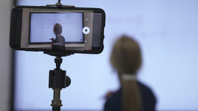 Portrait of very animated woman using an interactive whiteboard in a classroom teaching to phone camera.