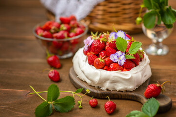 Pavlova dessert with strawberries and violet flowers as decor on a wooden background next to a plate full of strawberries