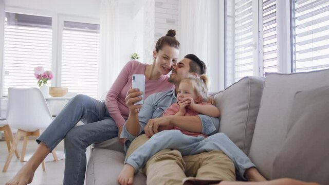 Young Family With Little Daughter Sitting On Sofa And Using Smartphone Indoors At Home