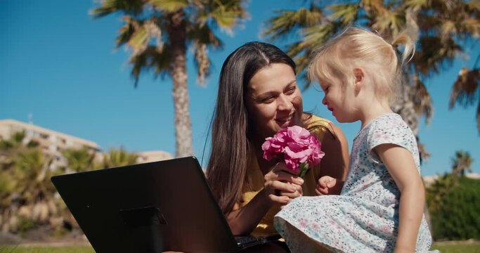 Young happy freelance woman working with laptop computer outdoors during baby walk. Cute child girl giving flowers ti her mother at public park in summer. 