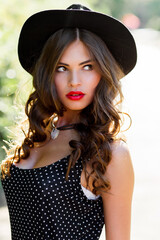 Summer fashion  portrait of r elegant woman with  perfect wavy  hair   in  stylish elegant black hat and bright make up posing in the park.  Street style.