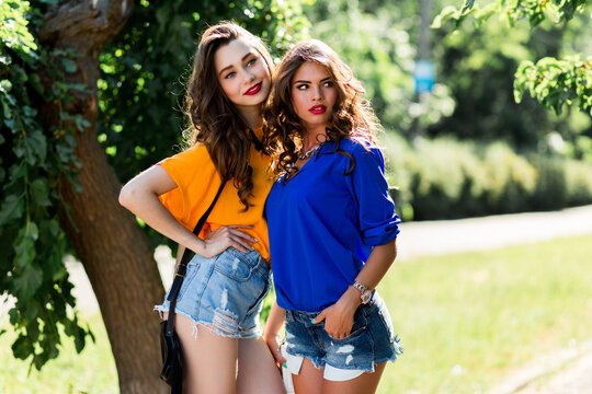 Two beautiful  girls walking in  the  summer park end talking.  Friends wearing stylish shirt and jeans shorts , enjoying  day off and have fun.