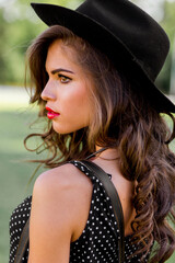 Summer fashion  portrait of r elegant woman with  perfect wavy  hair   in  stylish elegant black hat and bright make up posing in the park.  Street style.