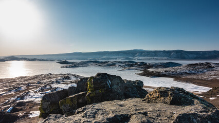 Fototapeta na wymiar Winter Siberian landscape. The sun is shining in the blue sky. Glare on the ice of a frozen lake. Picturesque rocks on a snow-covered ground. Baikal