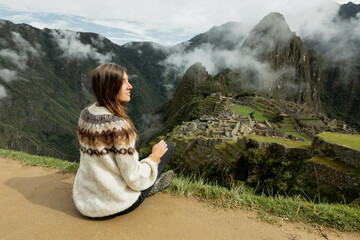 A young woman in a traditional Alpaca sweater sitting on the edge of Machu Picchu terrace