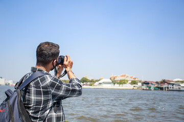 A South Asian tourist, wearing a striped shirt, picked up a camera to photograph Wat Arun on the ferry. and a backpack to walk happily during the long holiday happily in Bangkok, Thailand