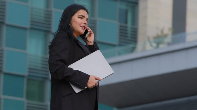 Successful young businesswoman walks down street against background of city building holds laptop confident hispanic girl manager talking by mobile phone answering business call speaking on cellphone