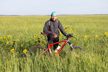 Young man with bicycle stands in the midst of field overgrown with sunflowers.