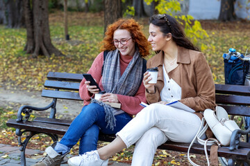 Two woman sitting at the bench in public park with coffee to go on hands and talk