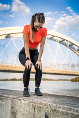 Young happy focused fitness girl in black yoga pants and orange short shirt work out and stretch her body on short concrete wall near river during the day.