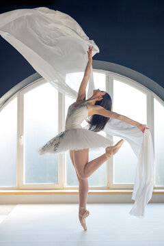 Young motivated ballerina dressed in white tutu costume practice ballet moves at ballet studio in front of big round shaped window background in natural ambient daylight atmosphere