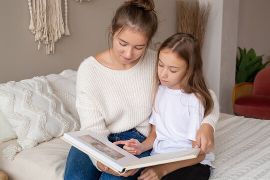 mother and daughter are sitting at home on the bed flipping through and looking at a book with photos. important moments of life in a photo album. Cute sisters read an interesting fairy tale together