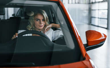 Sitting in the car. Woman in formal clothes is indoors in the autosalon