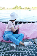 Rear view of happy young asian woman lay on pink pillow on net chair above river in cafe with copy space, relax time.