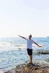 Fototapeta na wymiar Young attractive man in sunglasses in a white t-shirt and shorts stands on the shore of the mediterranean sea