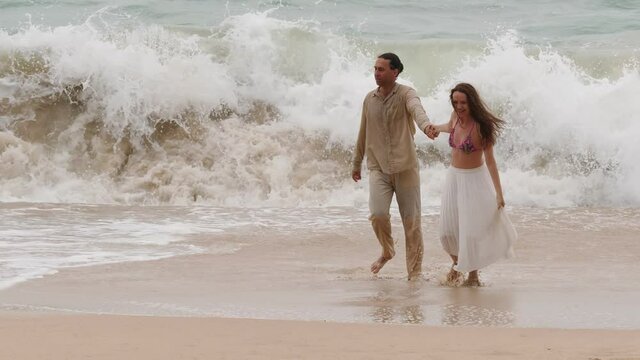Woman pull man by hand, away from growing wave, but guy walk lazily. Scary turbulent water crash down and loose strength, small uprush move on beach towards fearless couple