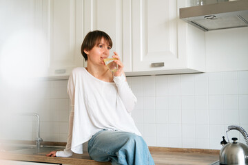 Young beautiful woman in casual clothes is sitting on countertop in kitchen and enjoying new day. Wellbeing, female health and good morning concept.