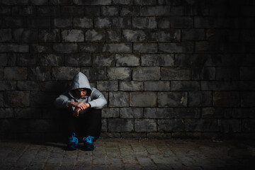 Sad bullied hispanic teenager sitting against wall in the darkness. Bullying in adolescence concept.