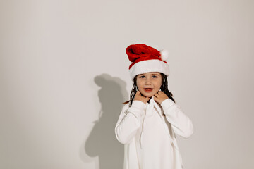Close up portrait of pretty happy little girl in white dress and Santa cap posing at camera over isolated background. Happy emotions, holiday mood