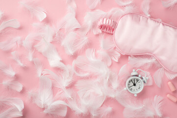 Top view photo of small white alarm clock light pink satin sleeping mask earplugs and pink feathers on isolated pastel pink background with copyspace