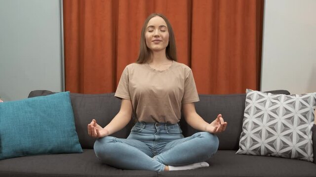 Pretty young woman is learing yoga at home. Smiling lady with closed eyes sitting in lotus position on couch at home. Relaxation, stress relief concept