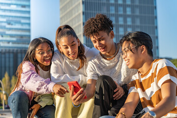 Group of happy teenage friends looking the phone and laughing in a bench in the city street.