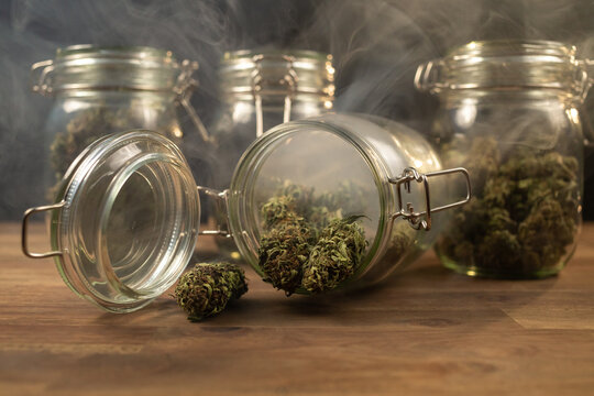 Cannabis buds spilled on the table from a storage glass jar, filled with smoke.