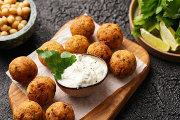 Traditional oriental chickpea deep fried falafel on a wooden board, tzatziki yoghurt sauce, hummus, fresh lime and green cilantro on black surface
