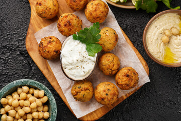 Traditional oriental chickpea deep fried falafel on a wooden board, tzatziki yoghurt sauce, hummus, lime and fresh green cilantro on black surface, top view