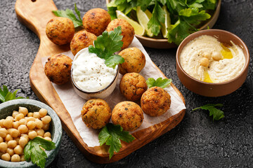 Traditional oriental chickpea deep fried falafel on a wooden board, tzatziki yoghurt sauce, hummus, fresh lime and green cilantro on black surface