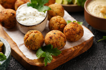 Traditional oriental chickpea deep fried falafel on a wooden board, tzatziki yoghurt sauce, hummus, fresh lime and green cilantro on black surface