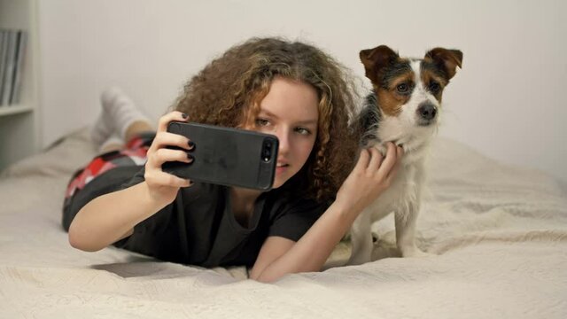 Beautifu teenage girl ltake a photo selfie bu mobile phone with her cute dog in cozy bedroom.