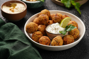 Traditional oriental chickpea deep fried falafel in a deep bowl with tzatziki yoghurt sauce, hummus, fresh lime and green cilantro on black surface