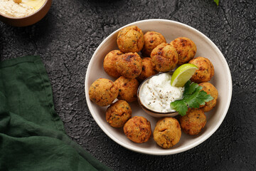 Traditional oriental chickpea deep fried falafel in a deep bowl with tzatziki yoghurt sauce, hummus, fresh lime and green cilantro on black surface