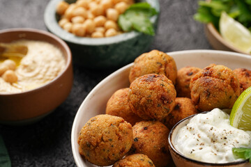 Traditional oriental chickpea deep fried falafel in a deep bowl with tzatziki yoghurt sauce, hummus, fresh lime and green cilantro on black surface