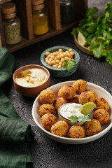 Traditional oriental chickpea deep fried falafel in a deep bowl with tzatziki yoghurt sauce, hummus, fresh lime and green cilantro on black surface