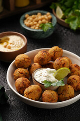 Traditional oriental chickpea deep fried falafel in a deep bowl with tzatziki yoghurt sauce, hummus, fresh lime and green cilantro on black surface