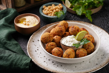 Traditional oriental chickpea deep fried falafel in a deep bowl with tzatziki yoghurt sauce, hummus, fresh lime and green cilantro on black surface