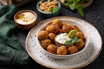 Traditional oriental chickpea deep fried falafel in a deep bowl with tzatziki yoghurt sauce, hummus, fresh lime and green cilantro on black surface