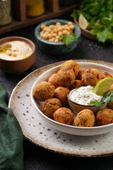 Traditional oriental chickpea deep fried falafel in a deep bowl with tzatziki yoghurt sauce, hummus, fresh lime and green cilantro on black surface