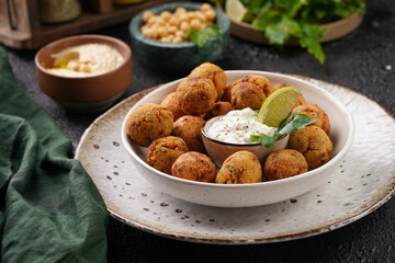 Traditional oriental chickpea deep fried falafel in a deep bowl with tzatziki yoghurt sauce, hummus, fresh lime and green cilantro on black surface