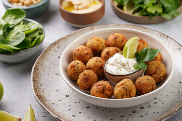 Traditional oriental chickpea deep fried falafel in a deep bowl with tzatziki yoghurt sauce, hummus, fresh lime and green cilantro on light grey concrete surface