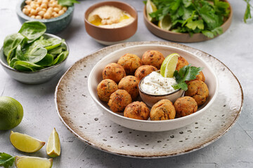 Traditional oriental chickpea deep fried falafel in a deep bowl with tzatziki yoghurt sauce, hummus, fresh lime and green cilantro on light grey concrete surface
