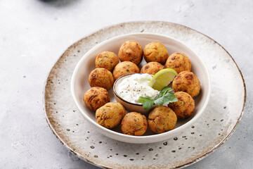 Traditional oriental chickpea deep fried falafel in a deep bowl with tzatziki yoghurt sauce, hummus, fresh lime and green cilantro on light grey concrete surface