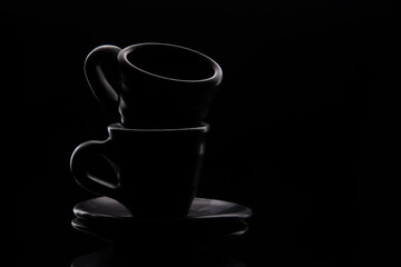 Two black ceramic coffee cups sit on top of each other and on saucers, against a black background with highlights and backlighting along the contour of the cups. Low key photography. Front view. 