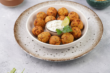 Traditional oriental chickpea deep fried falafel in a deep bowl with tzatziki yoghurt sauce, hummus, fresh lime and green cilantro on light grey concrete surface