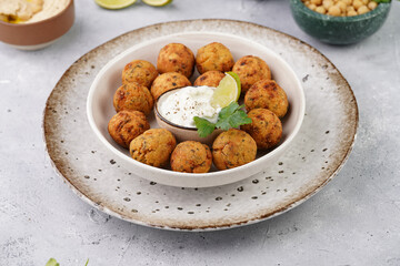 Traditional oriental chickpea deep fried falafel in a deep bowl with tzatziki yoghurt sauce, hummus, fresh lime and green cilantro on light grey concrete surface